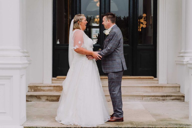 A smiling bride and groom facing each other on the steps of Winslade Manor. Both smiling, the groom has his hands on his bride's waist, as she holds his forearms