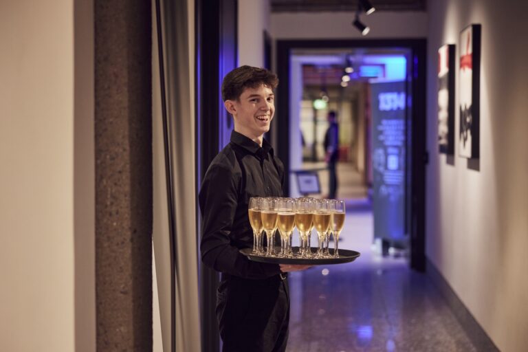 A smiling member of the Winslade Manor Events Team holding a tray of champagne flutes at an event in The Forge